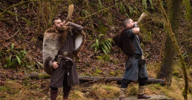 Two viking men playing the blow horn in a forest.