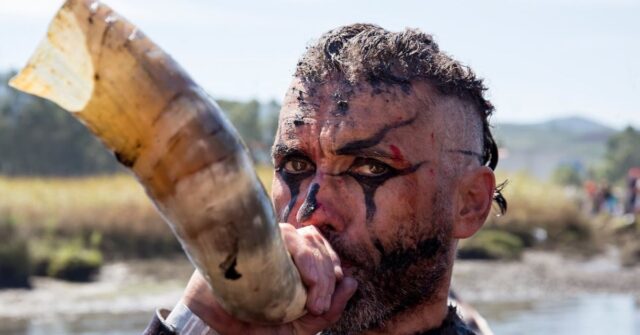 An intense looking viking warrior blowing a blow horn.