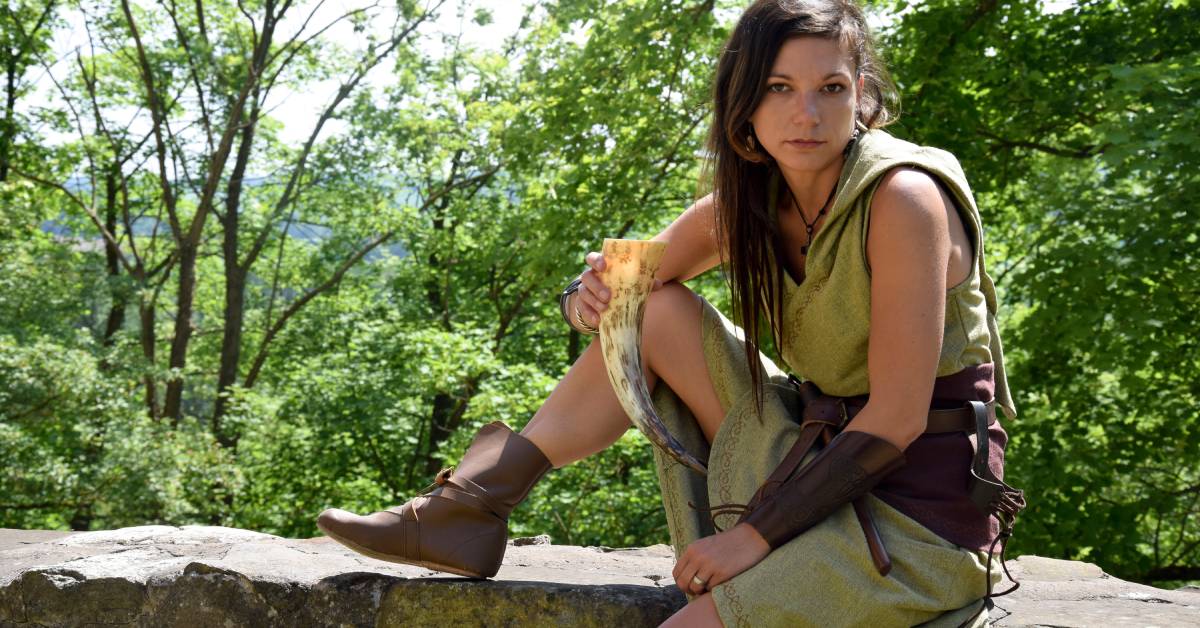 A nordic woman sitting on a wall while drinking from a horn.